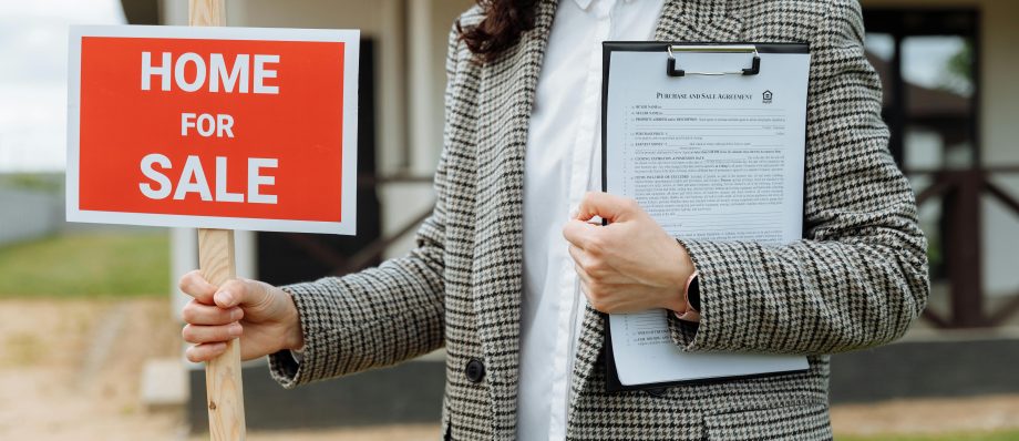 Why Do You Need an Estate Agent to Sell Your House? A Q&A Guide 2 A real estate agent holding a home for sale sign and clipboard outside a property.